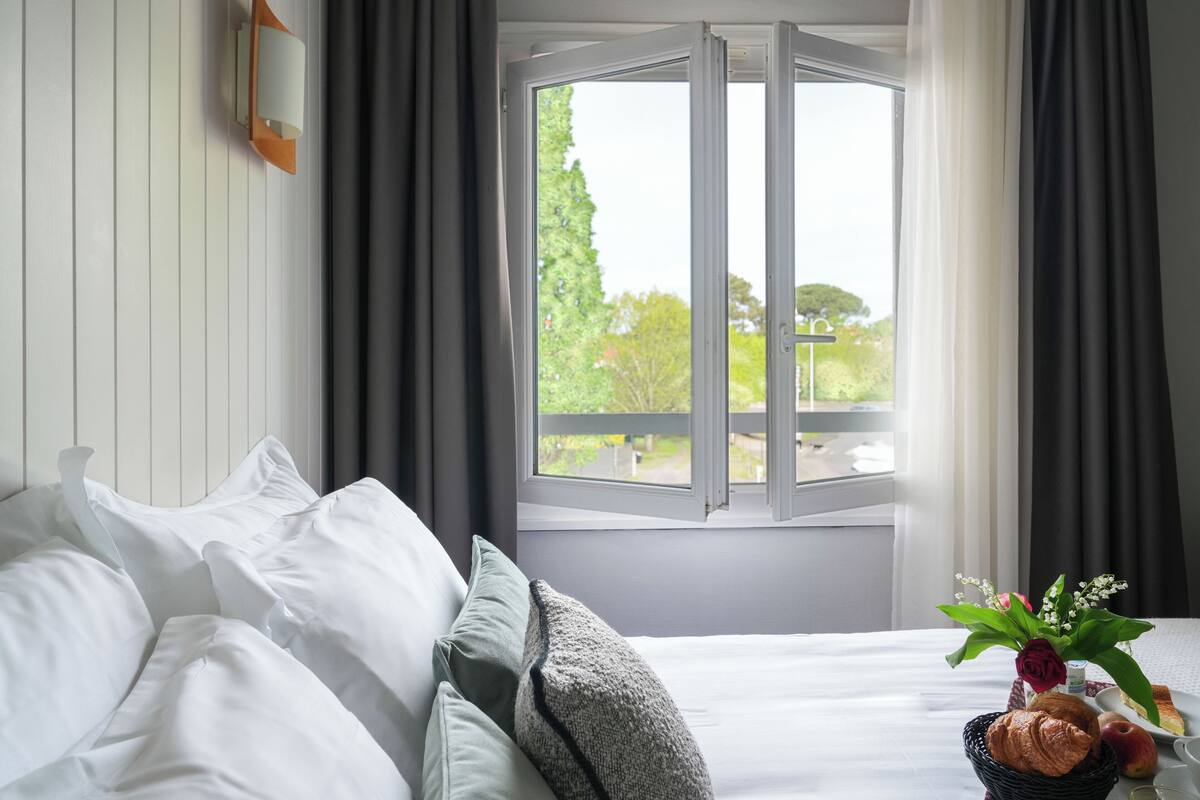 A neatly made bed with white linens, a window with curtains, and a small table with a basket containing a croissant, an apple, and a vase with flowers.