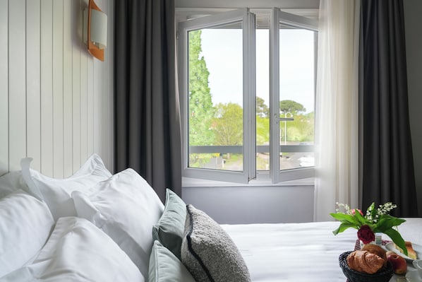 A neatly made bed with white linens, a window with curtains, and a small table with a basket containing a croissant, an apple, and a vase with flowers.