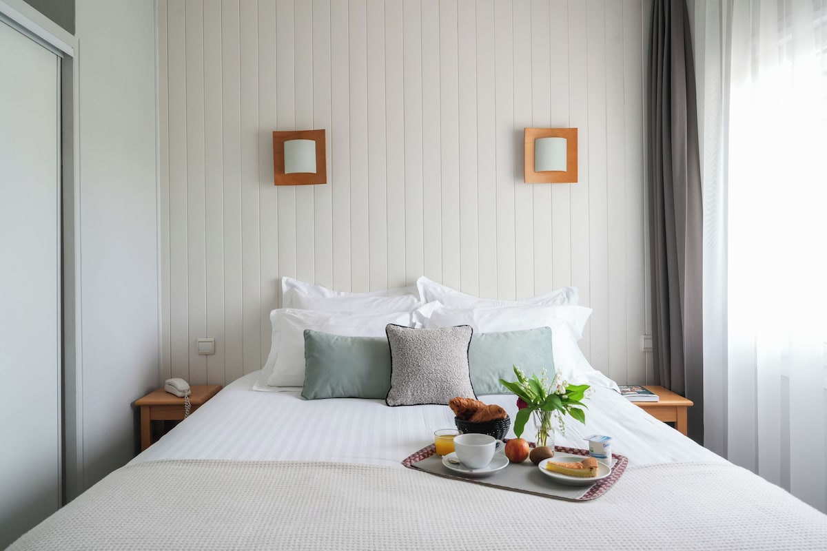 A neatly made bed with pillows, a bedside table with a phone, a tray with breakfast items, and a window with curtains.