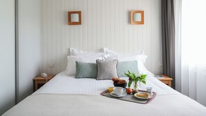Una cama bien hecha con almohadas, una mesita de noche con un teléfono, una bandeja con alimentos para el desayuno y una ventana con cortinas.