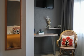 A room with a bed, a desk, a chair with a floral pillow, a mirror, a television, and a vase with dried flowers. - Hôtel Le Relais (Biscarrosse)