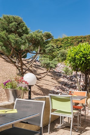 Un espace extérieur aménagé avec des chaises et des tables en métal, entouré de verdure et bordé d’une colline rocheuse.