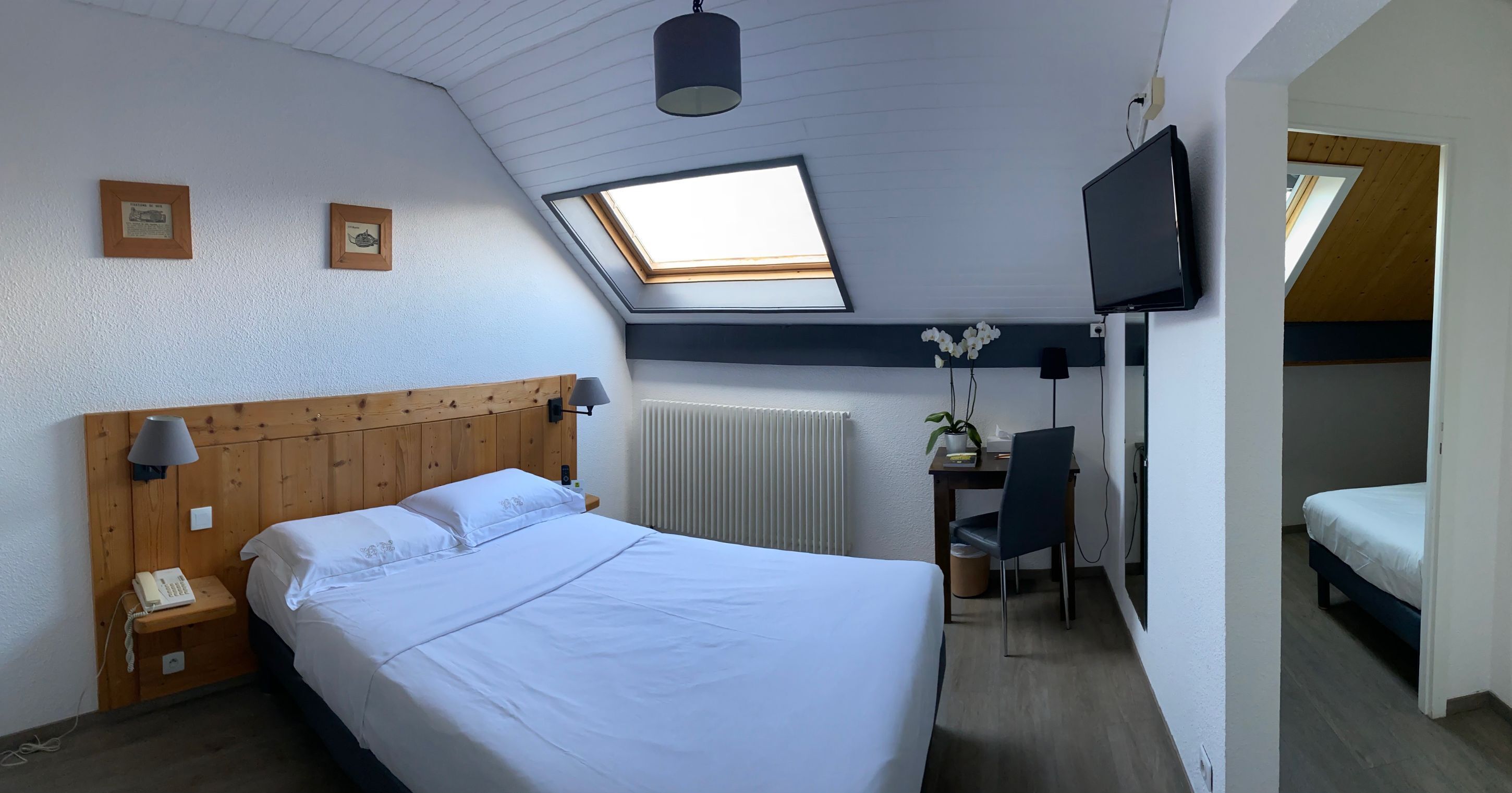A bedroom with a large bed, a desk with a chair, a TV mounted on the wall, and a skylight.