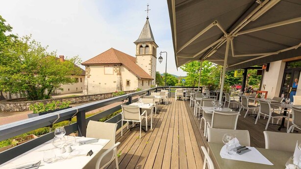 An outdoor dining area with tables and chairs, a view of a church with a steeple, and a paved walkway. - Logis Hôtel Annecy Nord / Argonay (Argonay)