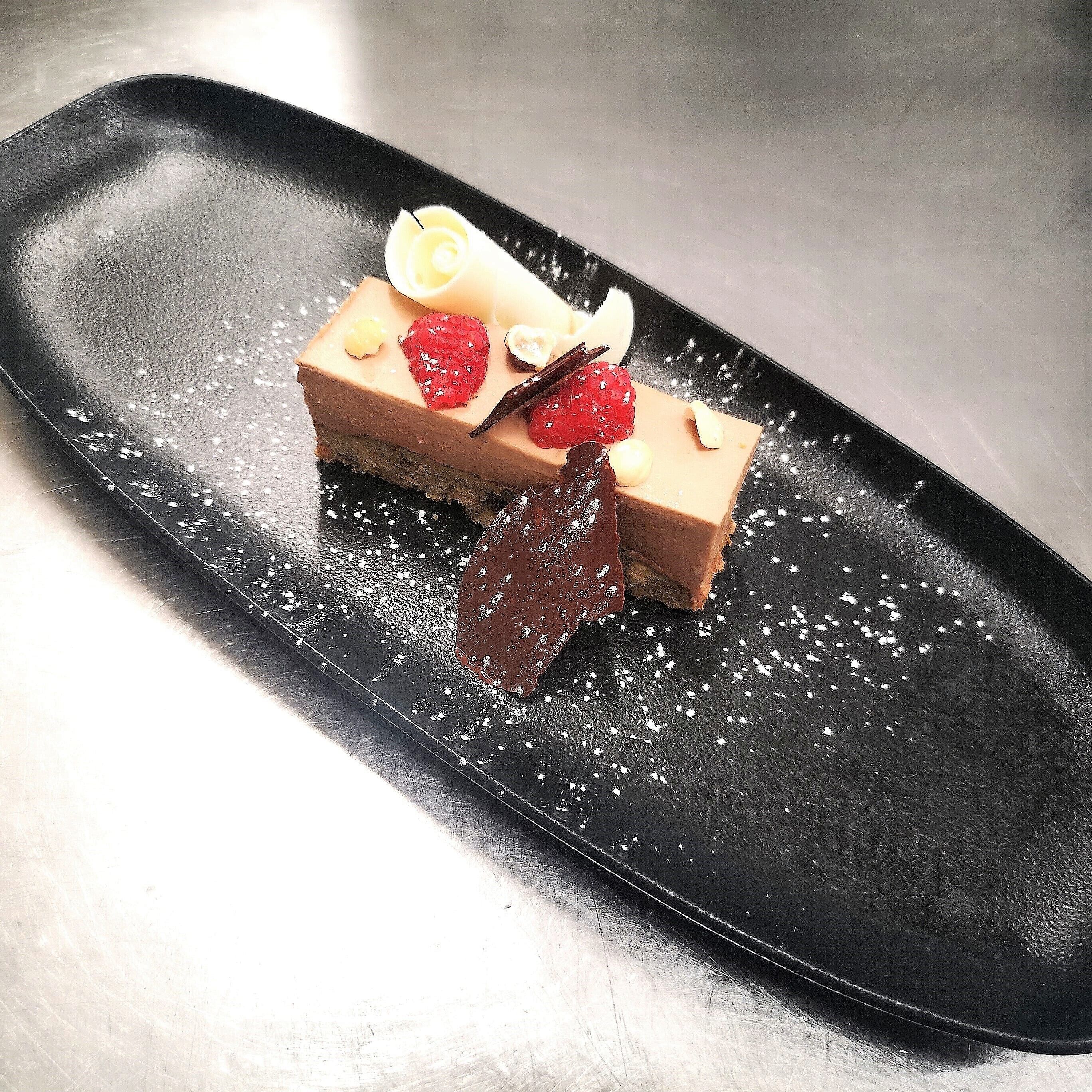 Postre sobre un plato negro, con azúcar glas, una hoja de chocolate y guarniciones de fruta.