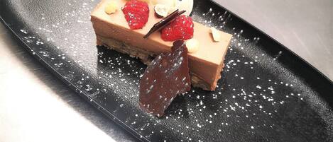 Postre sobre un plato negro, con azúcar glas, una hoja de chocolate y guarniciones de fruta.