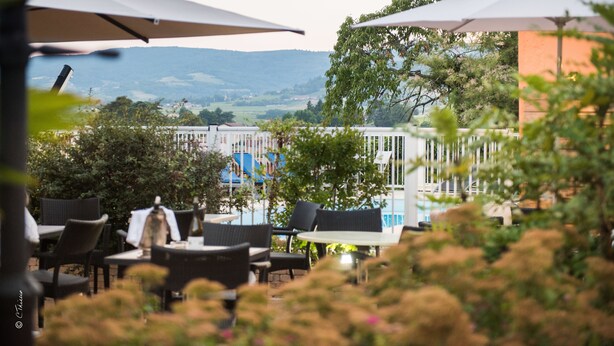 An outdoor seating area with tables and chairs, umbrellas, and a view of buildings and hills in the background. - Chez La Rose (Juliénas)