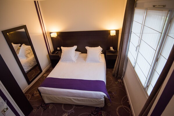 A hotel room with a large bed, a bedside table with a lamp, a mirror, and a window with curtains.