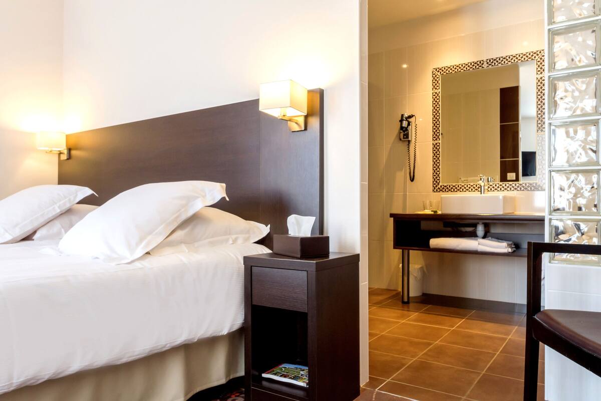 A hotel room with a bed, bedside lamp, nightstand, and a bathroom with a mirror, sink, and towel rack.