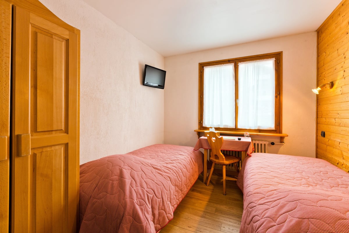 A small room with two single beds, a desk with a chair, a television, and a window with curtains.