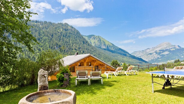A wooden cabin with outdoor seating and a ping pong table, surrounded by lush greenery and mountains. - Le Roitelet (Chatel)