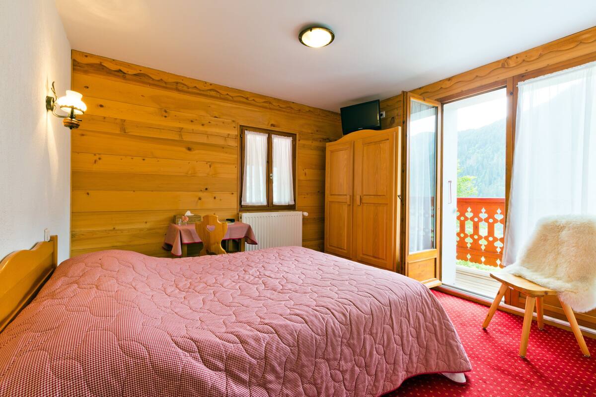 A bedroom with a large bed, wooden walls, a small table with chairs, and a view of a balcony.