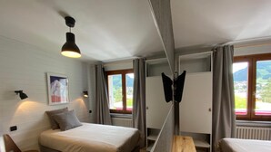 A bedroom with a bed, a wooden bench, a chair, a hanging light fixture, and a window with curtains. - Le Roitelet (Chatel)