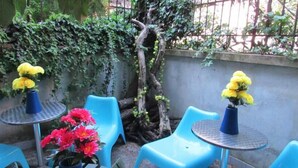 Un patio con sillas y mesas de plástico azul, rodeado de follaje y un tronco de árbol.