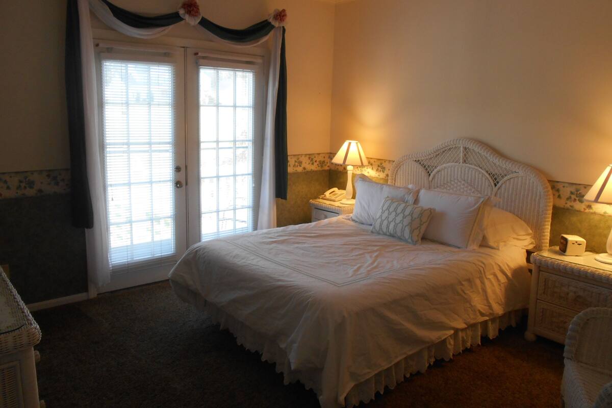 A bedroom with a large bed, two bedside tables with lamps, a wicker headboard, and a door with curtains.