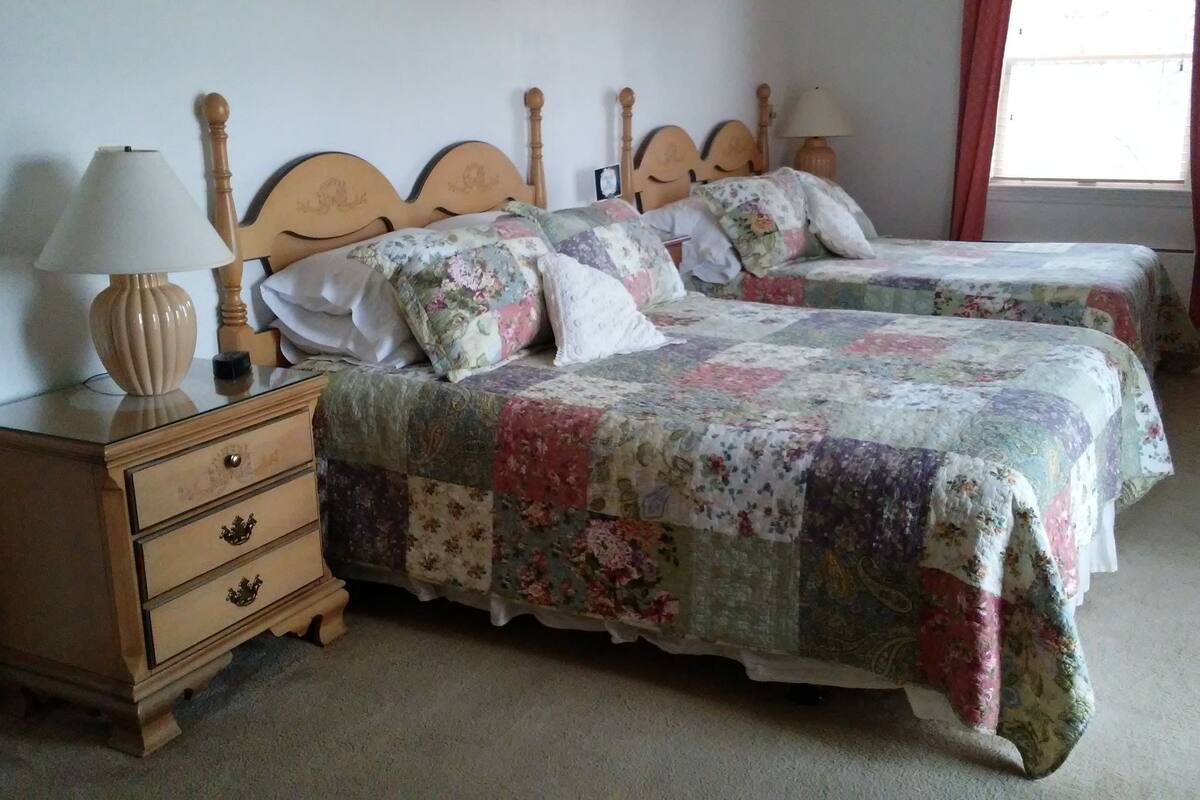 A bedroom with two wooden beds, each with a floral quilt, a bedside table with a lamp, and a wooden dresser with drawers.