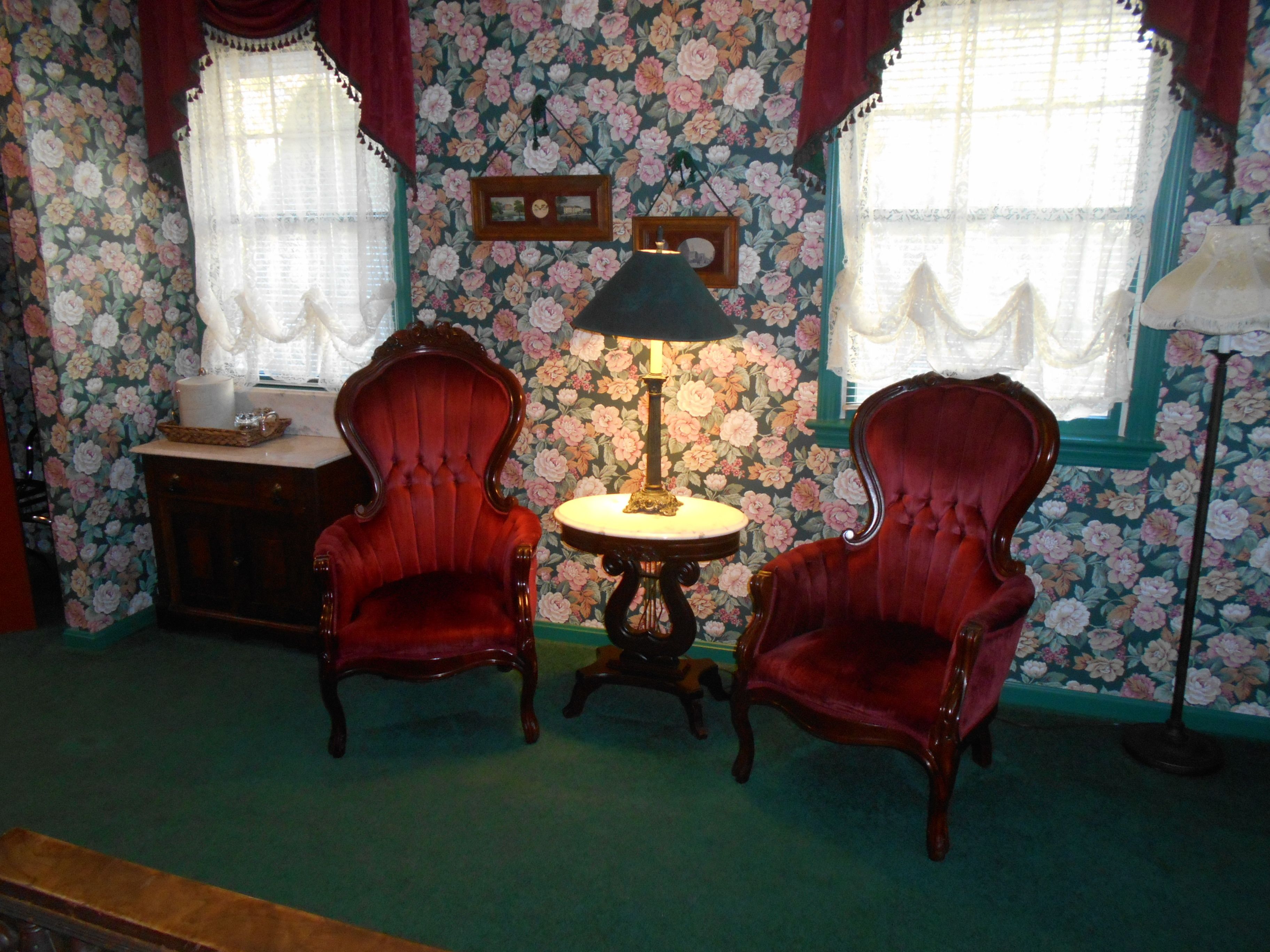 A room with floral wallpaper, two red armchairs, a small table with a lamp, and a dresser.
