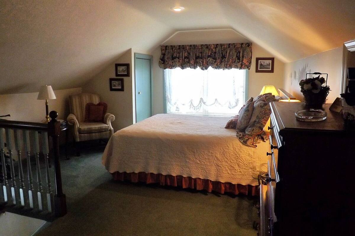 A bedroom with a large bed, a chair, a desk, and a window with curtains.