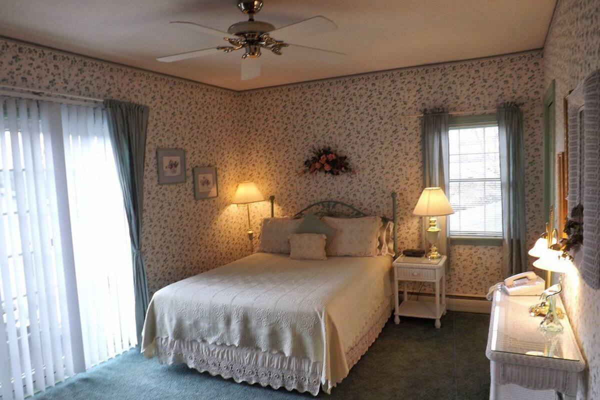A bedroom with a bed, two bedside lamps, a nightstand, a dresser, a ceiling fan, and a window with curtains.