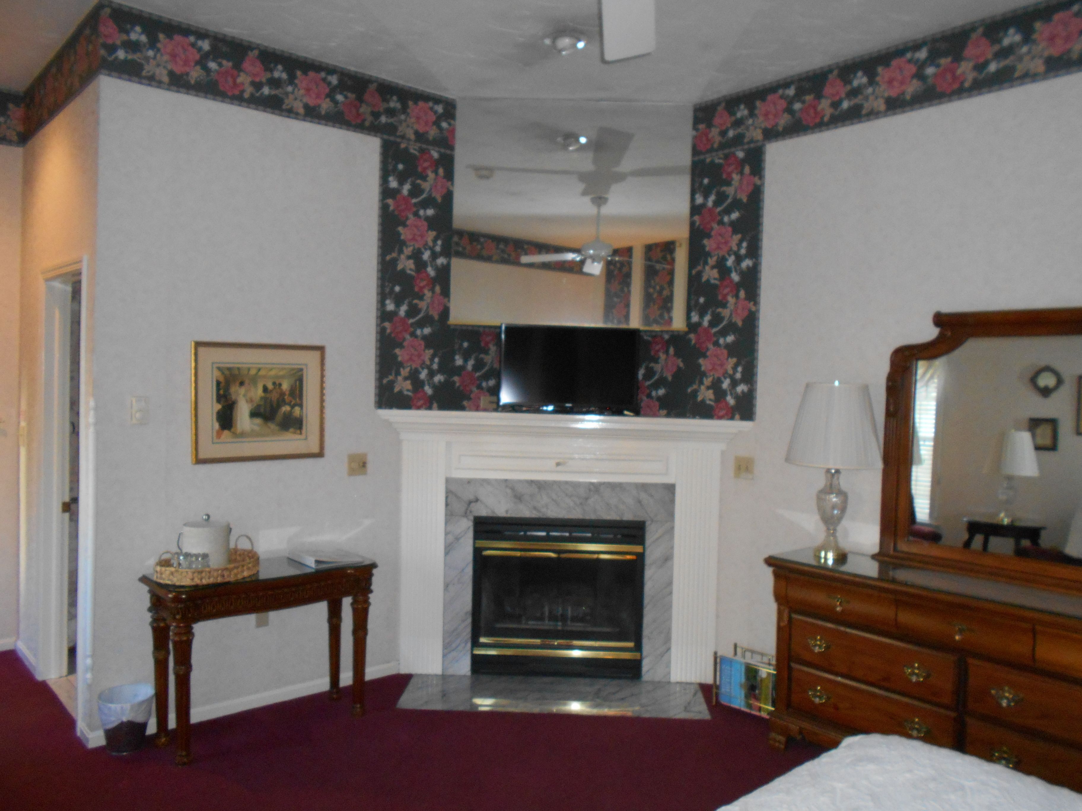 A hotel room with a fireplace, a television above it, a wooden dresser, and a framed picture on the wall.