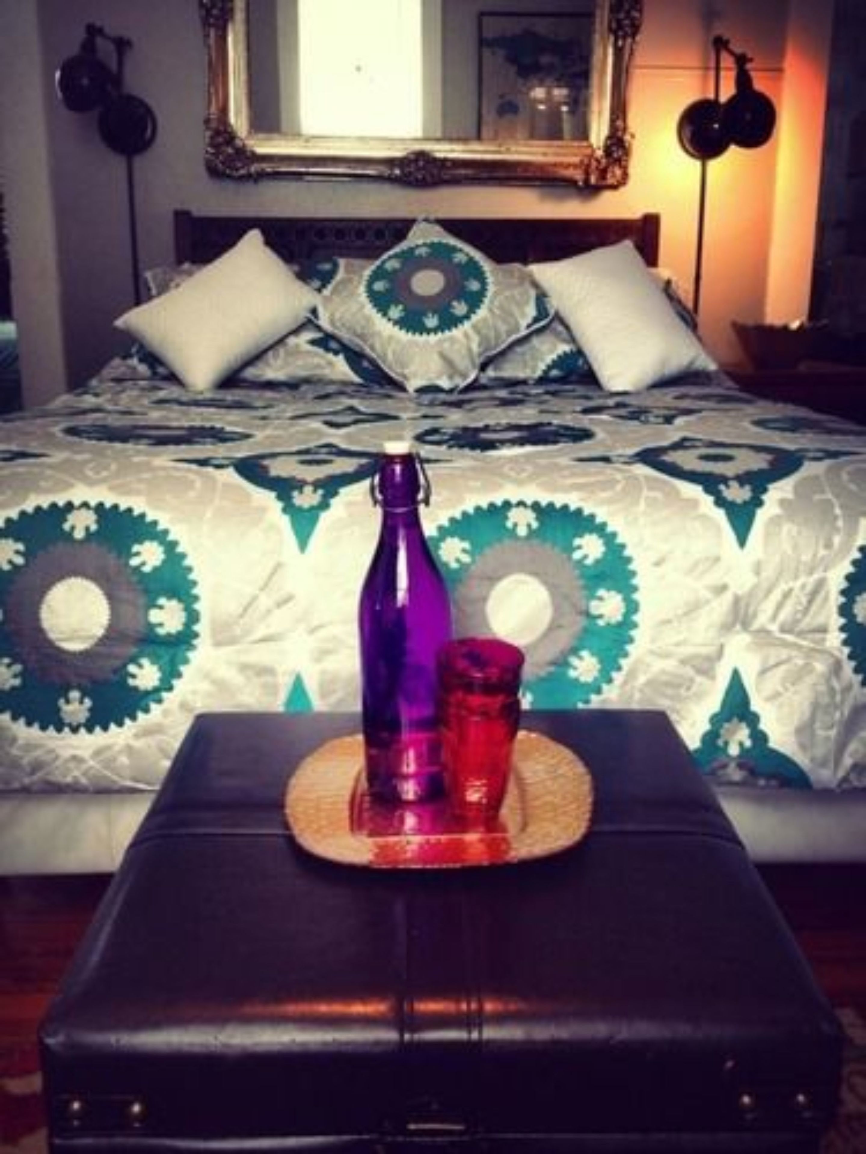 A bed with patterned bedding, a bedside table with two bottles, and a mirror above the bed.