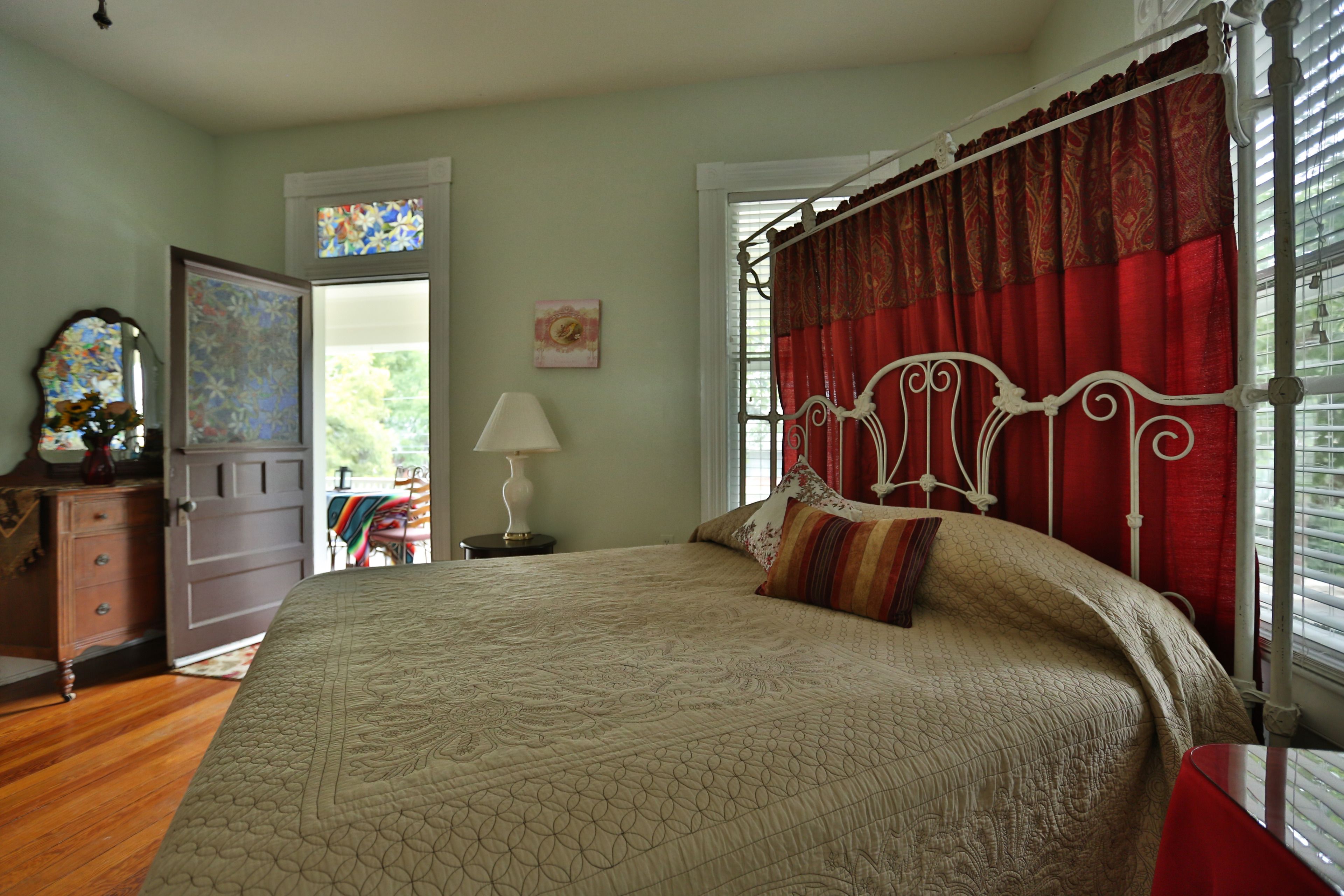 A bedroom with a large bed, a wooden dresser, a nightstand with a lamp, a window with red curtains, and a door leading to another room.