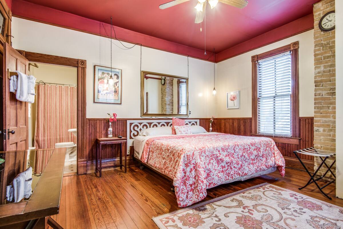 A bedroom with a wooden floor, a bed with a floral bedspread, a wooden headboard, a nightstand, a mirror, a window with blinds, and a clock on the wall.