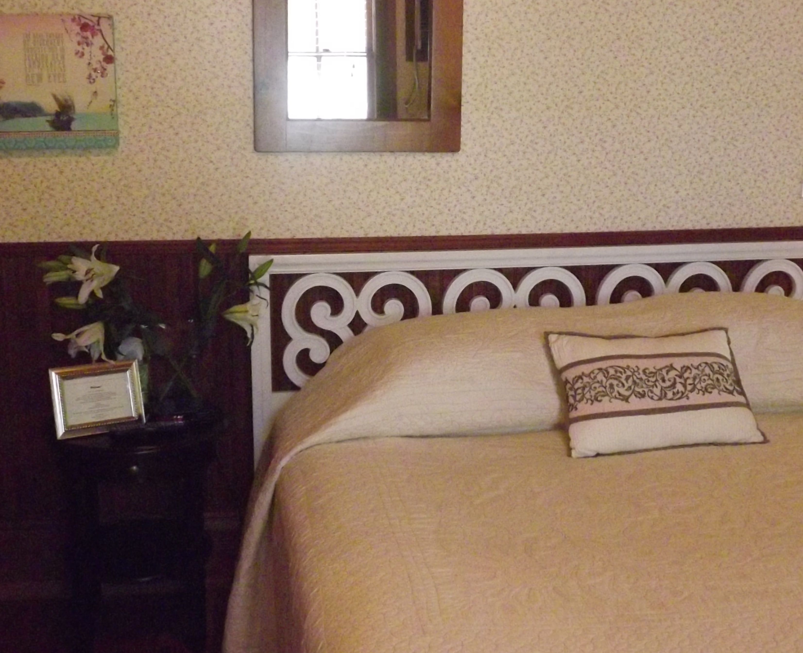 A hotel room with a neatly made bed, a bedside table with a vase of flowers, and a framed certificate on the wall.