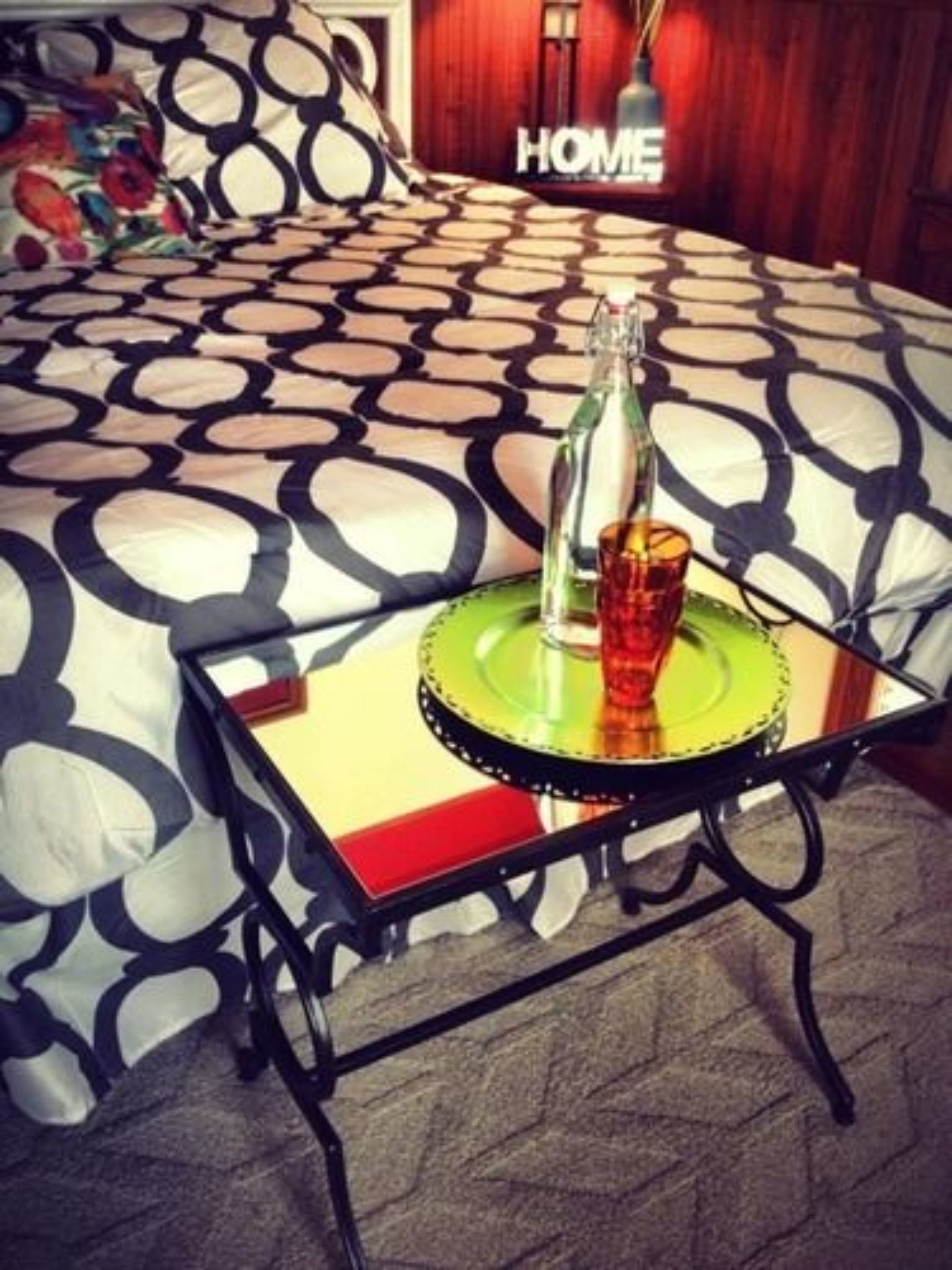 A bed with a patterned bedspread, a bedside table with a bottle and glass, and a 'HOME' sign in the background.
