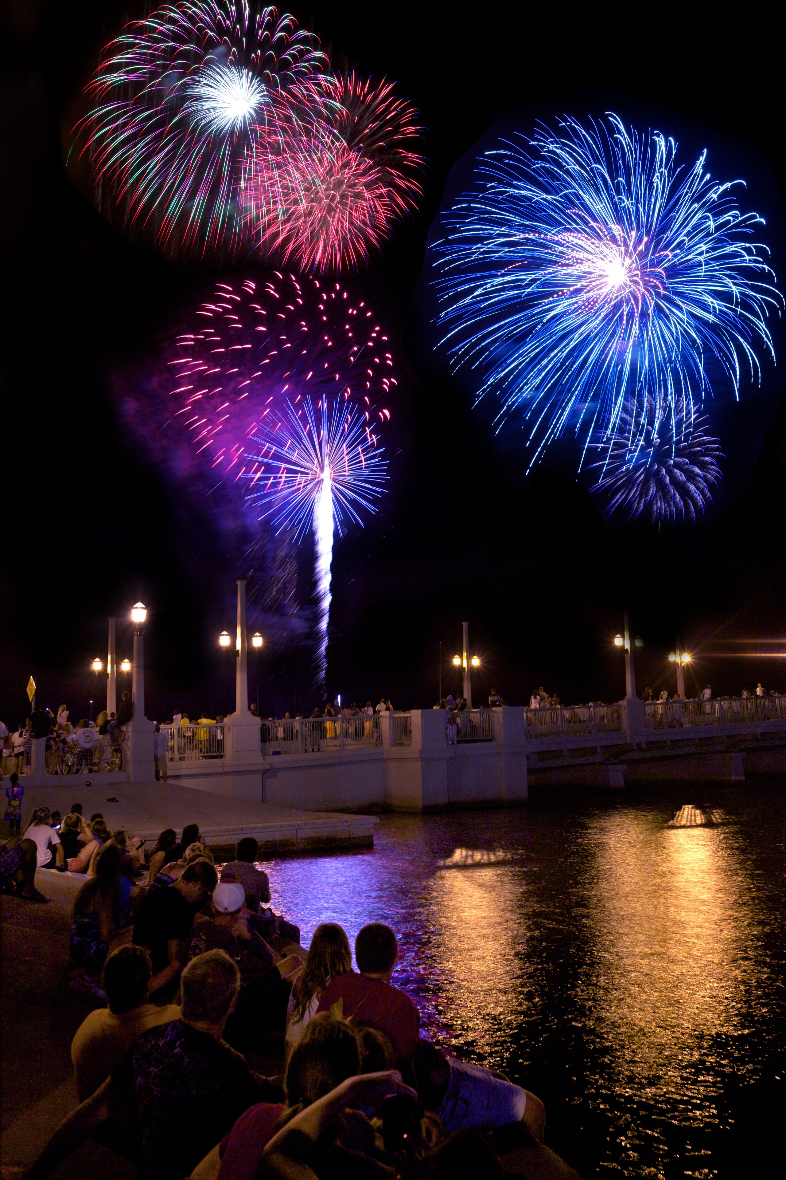 A waterfront view with a crowd of people watching fireworks.
