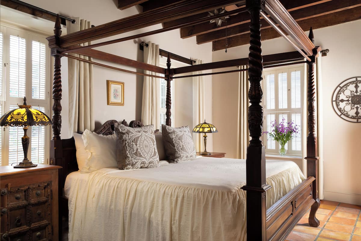 A four-poster bed with a wooden frame, lace pillows, and a cream-colored bedspread. There is a wooden nightstand with a lamp and a dresser with drawers. A vase with flowers is on the dresser, and a wall clock is on the wall.