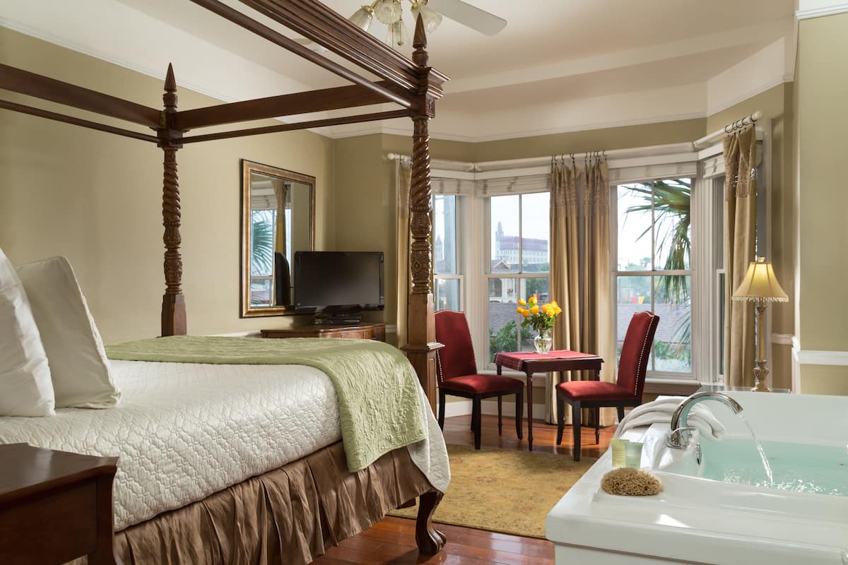 A four-poster bed with a green bedspread, a television on a wooden nightstand, a red chair, a small table with a vase of flowers, and a bathtub with a showerhead.