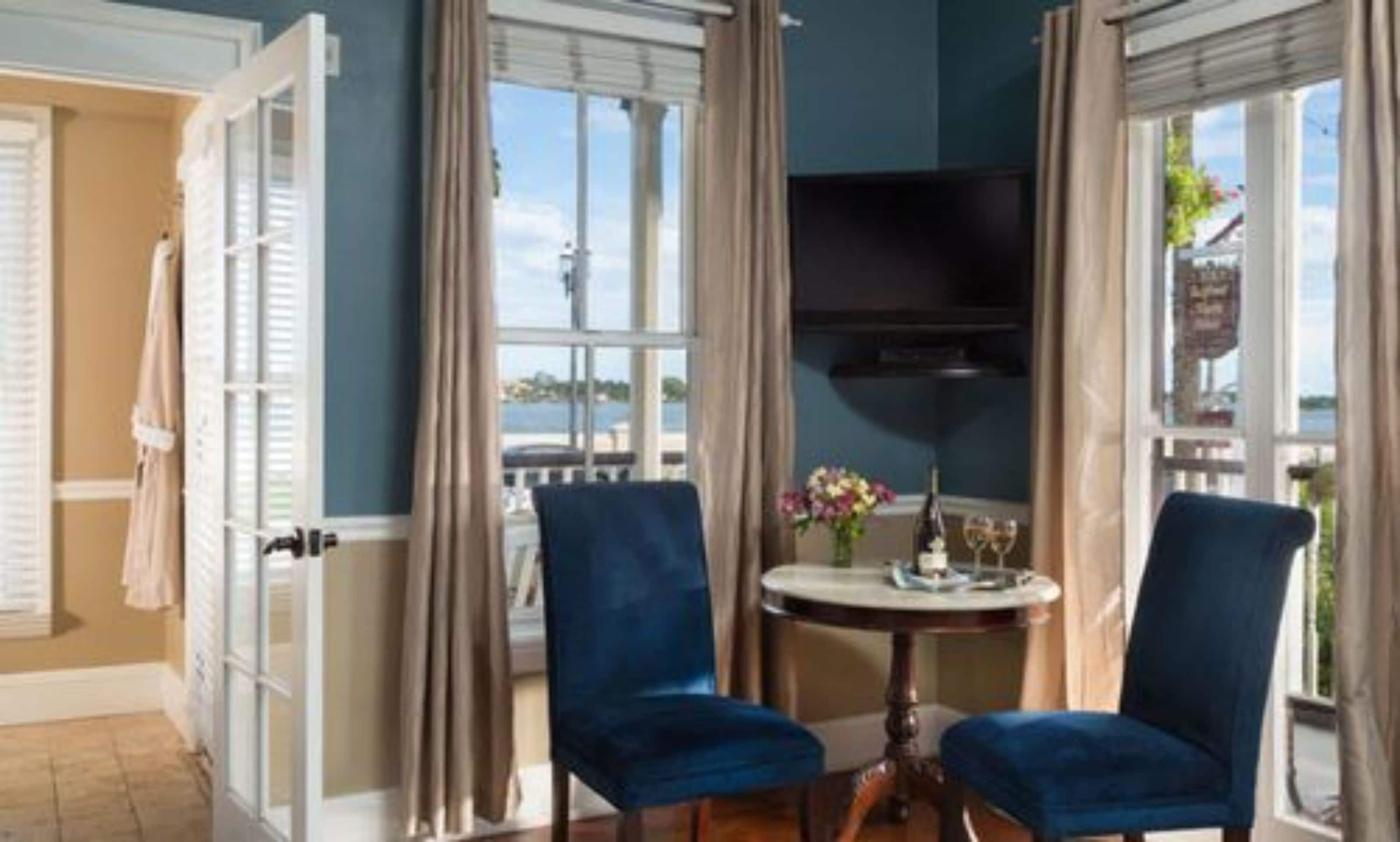A room with two blue chairs, a small round table with a vase of flowers, and a television mounted on the wall.