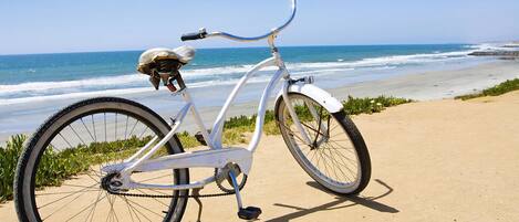 A white bicycle with a helmet on the seat, parked on a sandy beach with the ocean in the background.