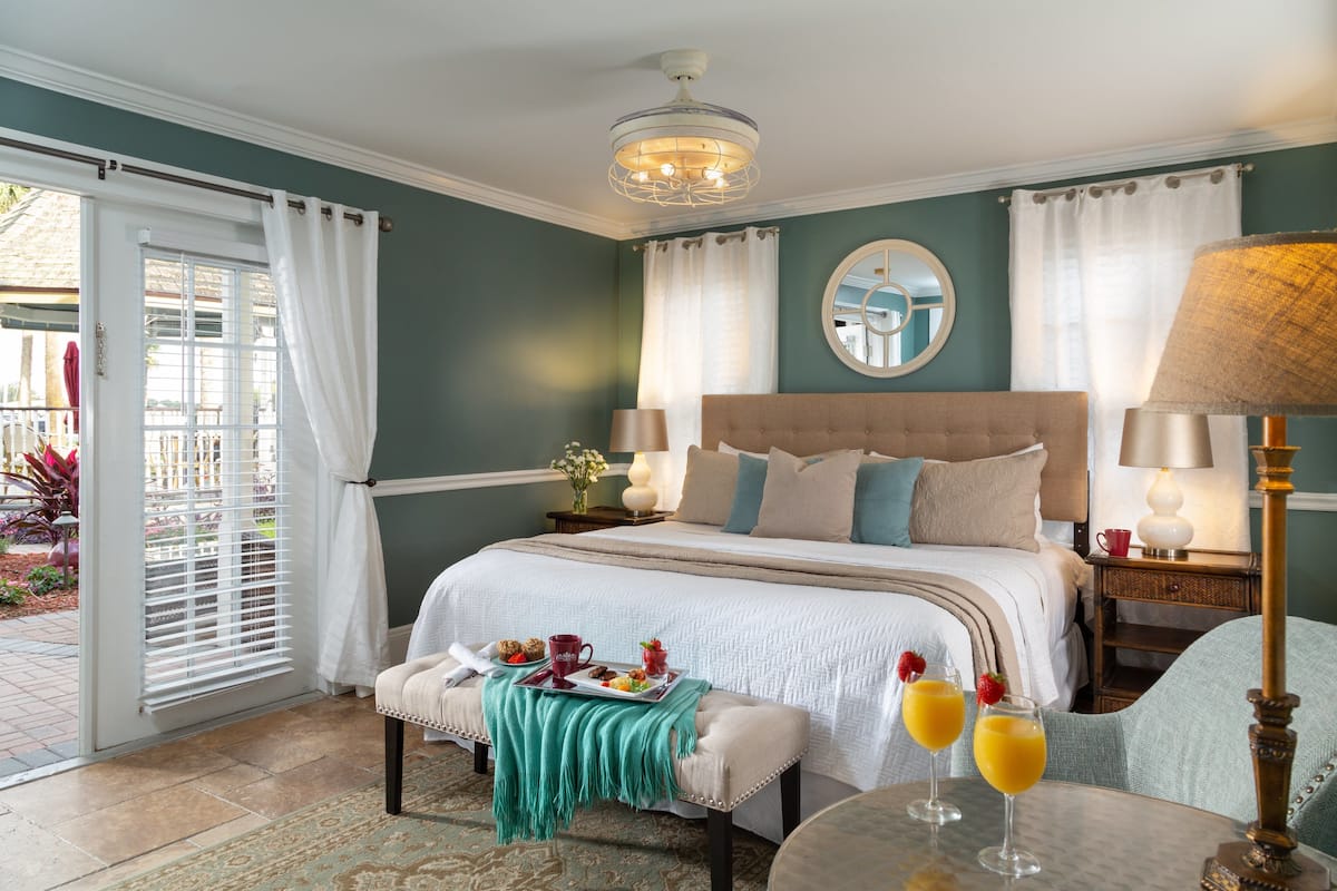 A bedroom with a large bed, a bench, a table with drinks, and a view of the outside.