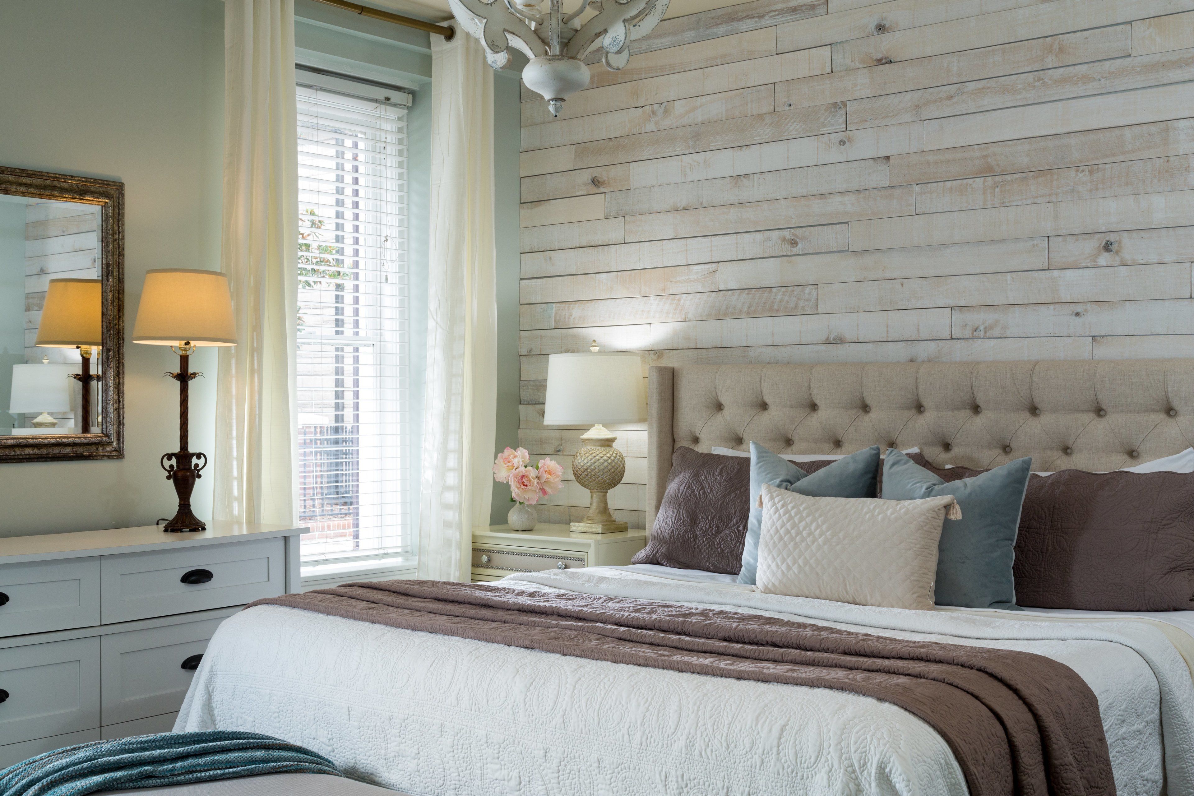 A bedroom with a wooden headboard, a large bed with multiple pillows, a nightstand with a lamp, and a dresser with drawers.