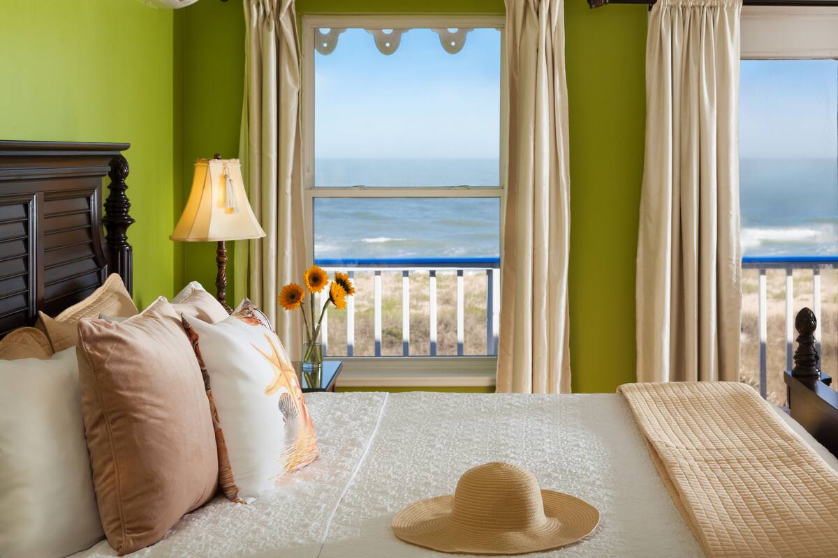 A bedroom with a bed, pillows, a hat, a lamp, and a view of the ocean through a balcony.