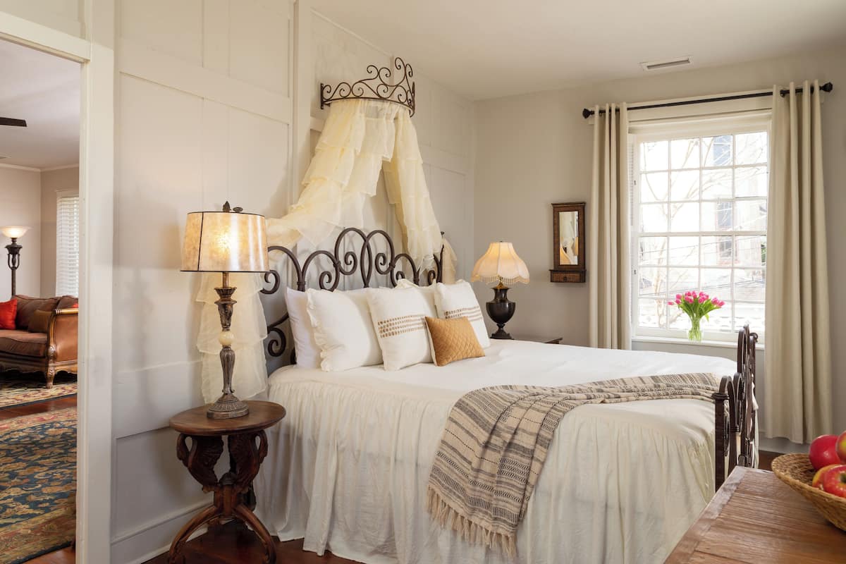 A bedroom with a canopy bed, a large window with curtains, a wooden floor, and a view of the outside.