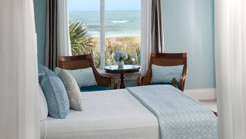 A bedroom with a bed, two chairs, a small table with a vase and glasses, and a view of the ocean.