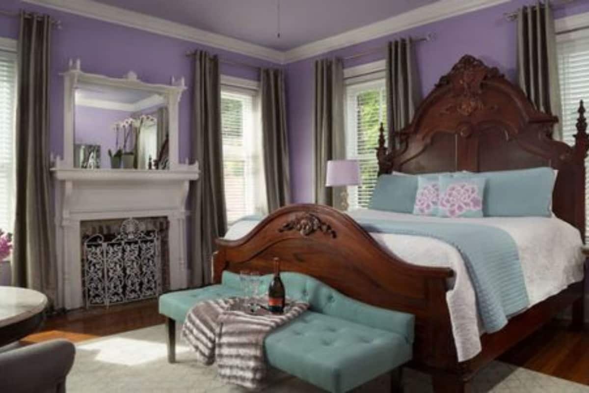 A bedroom with a large bed, a fireplace, a window with curtains, and a small table with a bottle.