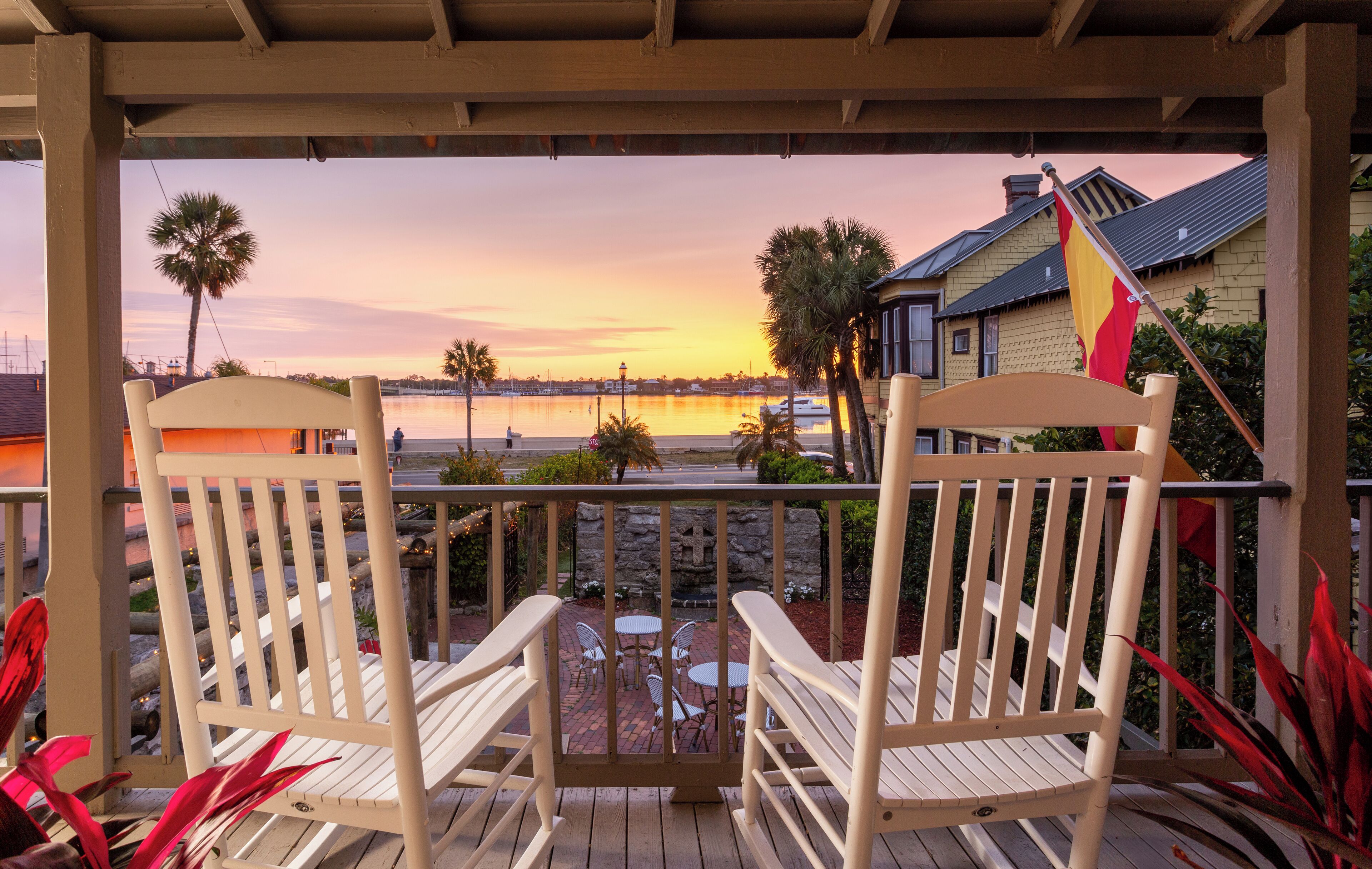 A wooden deck with two rocking chairs and a small table, overlooking a body of water and a sunset.