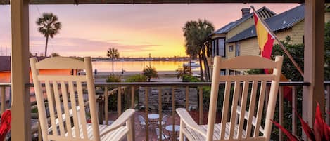 A wooden deck with two rocking chairs and a small table, overlooking a body of water and a sunset.