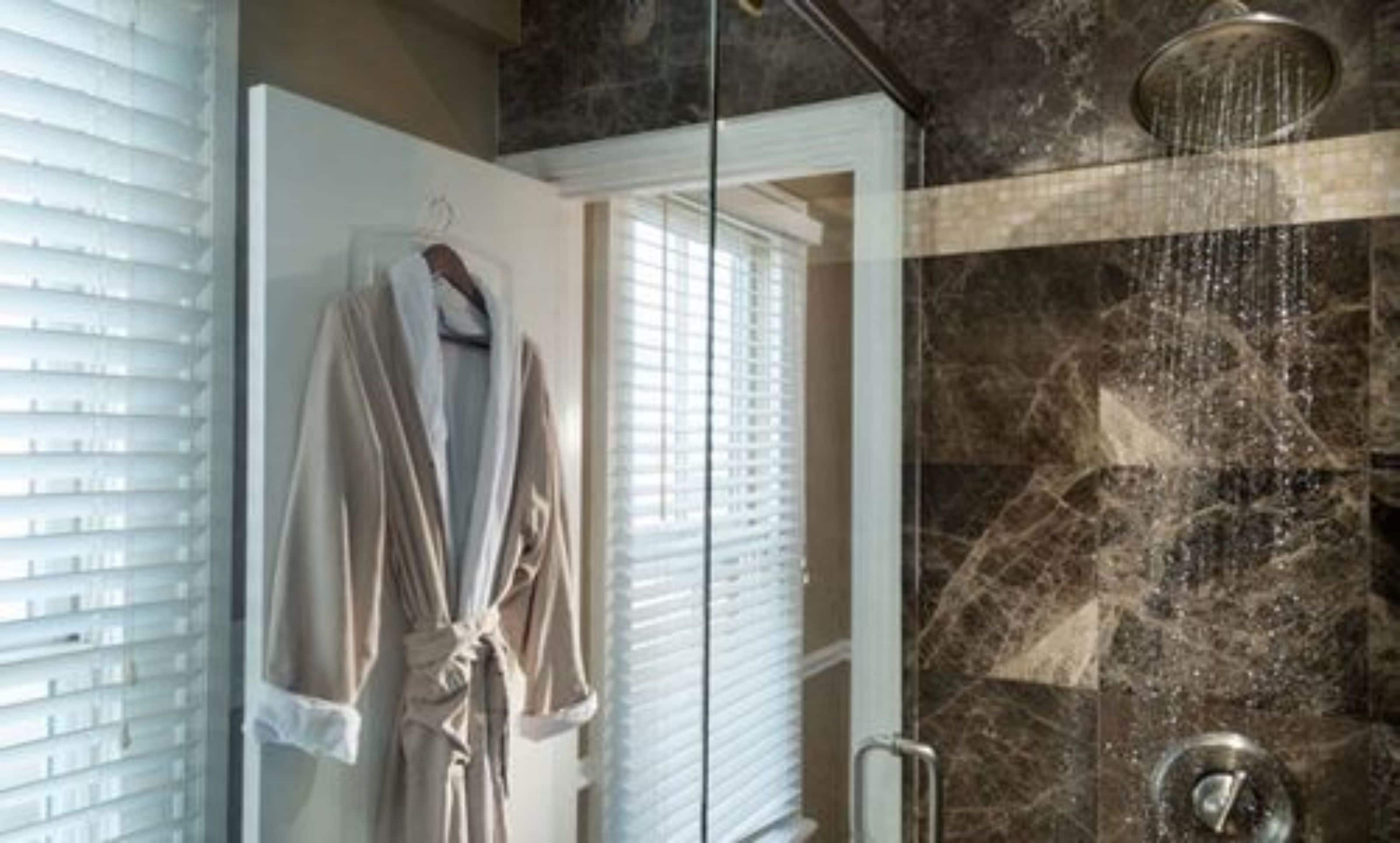 A bathroom with a glass shower enclosure, a robe hanging on the wall, and a window with blinds.