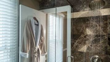 A bathroom with a glass shower enclosure, a robe hanging on the wall, and a window with blinds.
