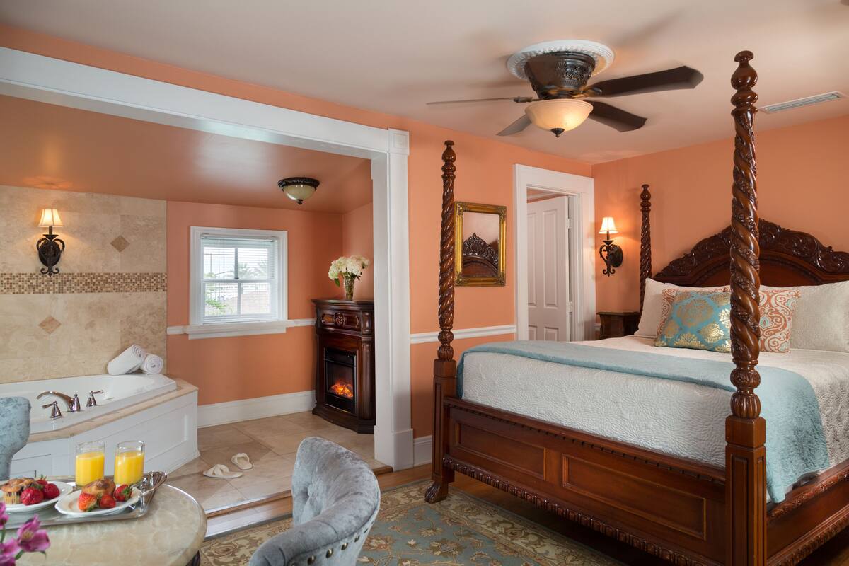 A bedroom with a four-poster bed, a sitting area with a chair, a fireplace, and a large window.