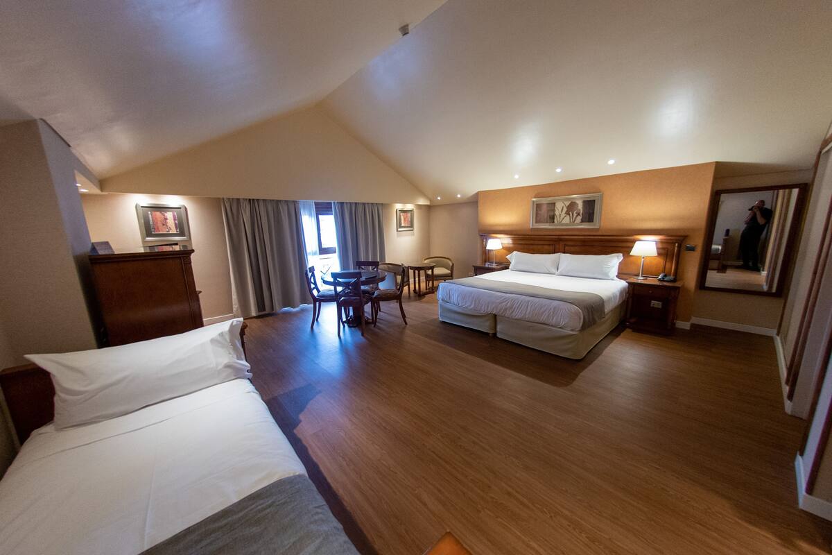 A hotel room with a large bed, a smaller bed, a desk with a chair, a wooden dresser, and a painting on the wall.