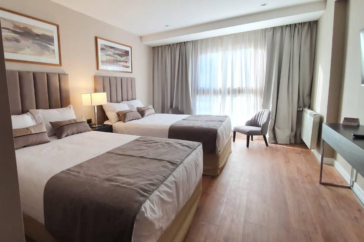 A hotel room with two beds, a desk, a chair, and a large window with curtains.