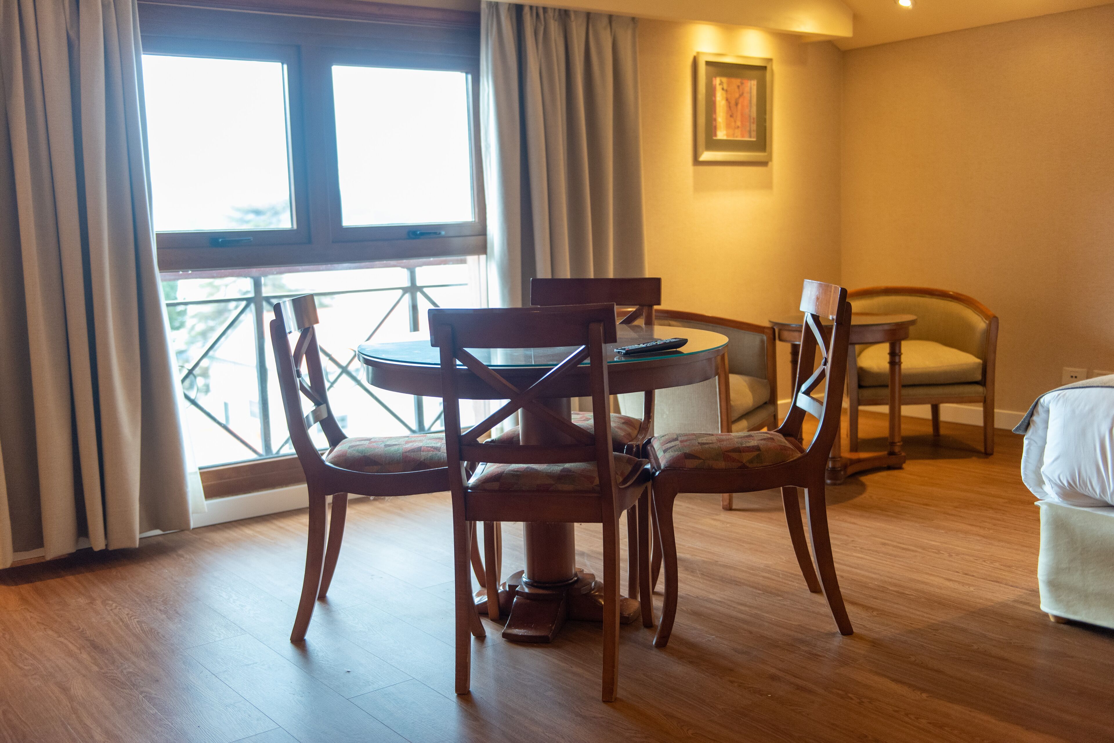 Habitación de hotel con una mesa de comedor de madera, cuatro sillas, una mesita redonda, un sofá y una ventana con cortinas.