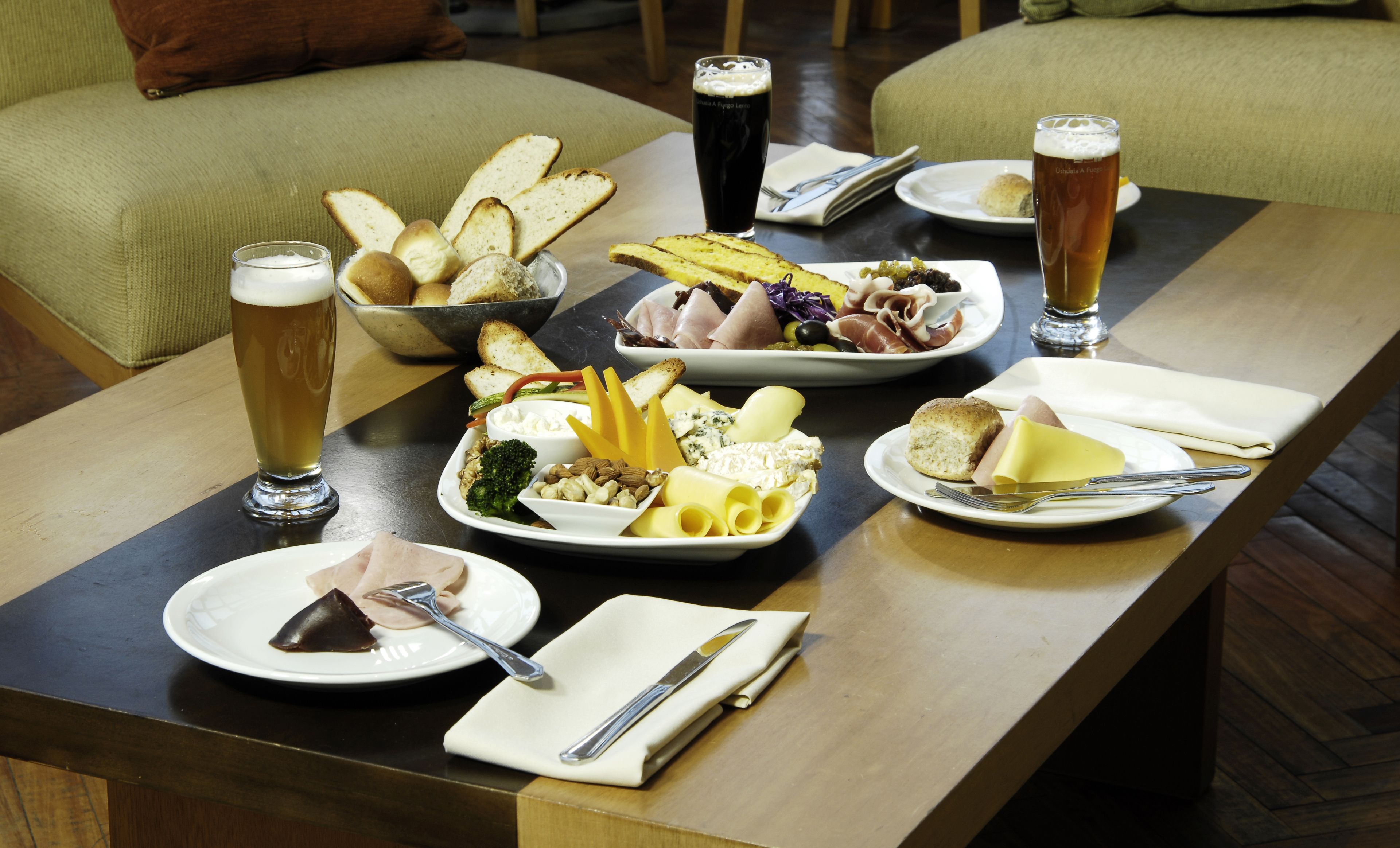 Una mesa de comedor preparada con diversos quesos, embutidos y pan, acompañada de vasos de cerveza.
