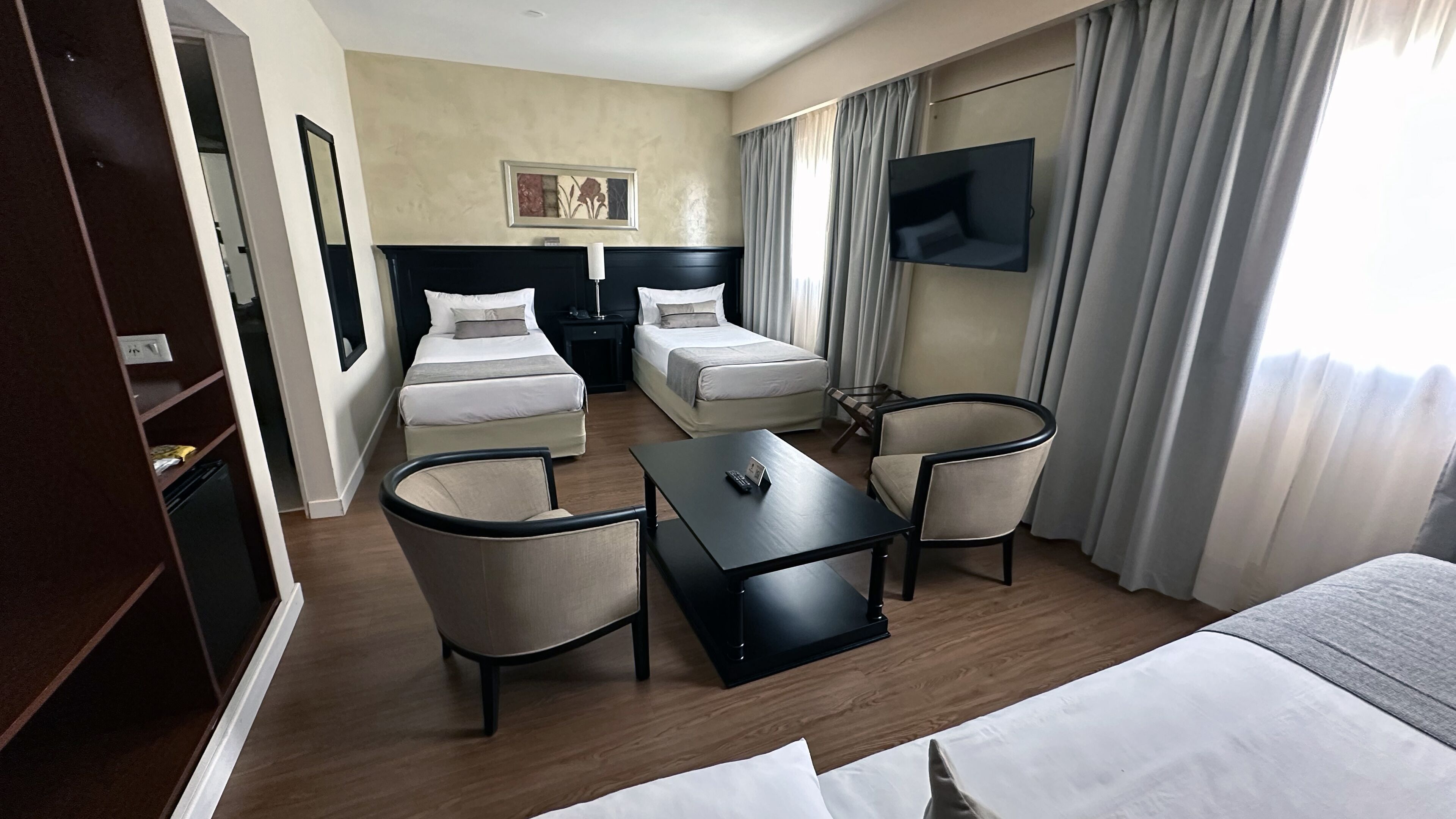 Habitación de hotel con cama doble, dos camas individuales, escritorio, dos sillas, televisor y ventana con cortinas.